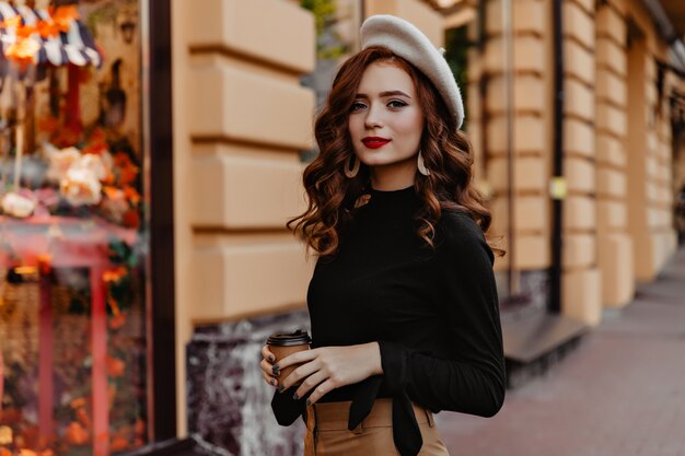 Magnífica chica europea en boina marrón pasar tiempo al aire libre. Curiosa dama de jengibre francés de pie en la calle y bebiendo té.
