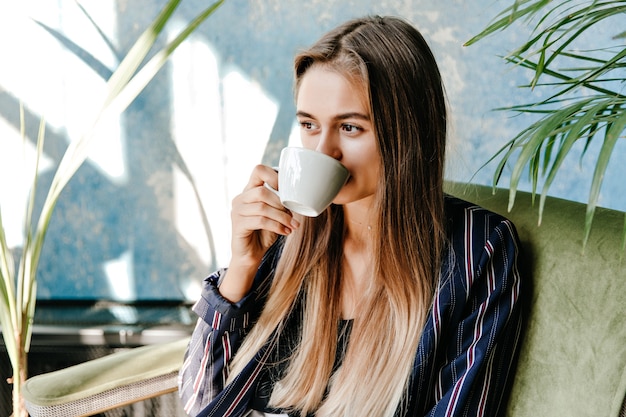 Magnífica chica de pelo largo tomando café con gusto. Atractiva modelo femenina disfrutando de la mañana en casa.