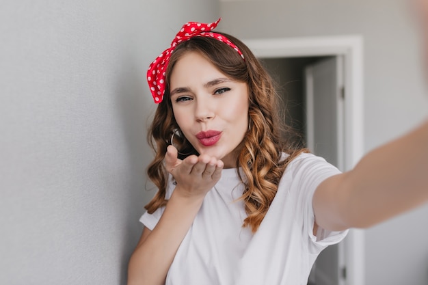 Magnífica chica rizada con maquillaje nude enviando beso al aire. Modelo femenino blanco guapo haciendo selfie.