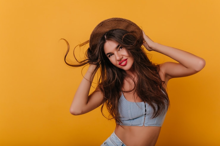 Foto gratuito magnífica dama bronceada bailando en el espacio amarillo y tocando su sombrero