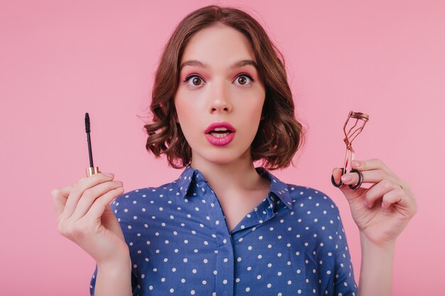 Magnífica dama con maquillaje rosa brillante posando con rímel. Foto interior de niña linda sorprendida riza sus pestañas.