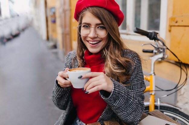 Magnífica dama de ojos azules en boina francesa disfrutando de un café en la cafetería al aire libre