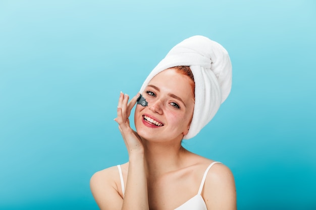 Magnífica mujer joven aplicando mascarilla. Foto de estudio de chica inspirada haciendo tratamiento de cuidado de la piel con sonrisa sobre fondo azul.