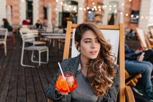 Magnífica mujer pálida con cabello castaño claro mirando a otro lado con expresión seria y bebiendo cócteles