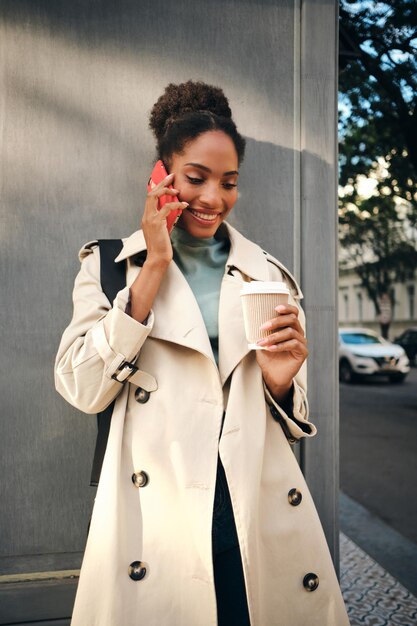 Magnífica niña afroamericana sonriente con elegante gabardina con café para ir felizmente hablando por celular al aire libre