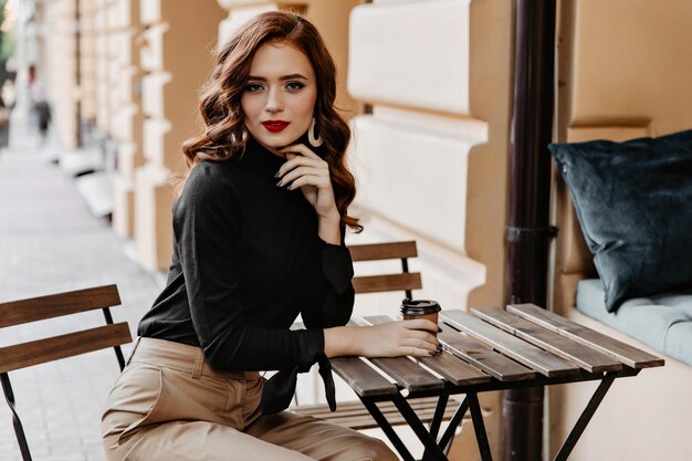 Magnífica señorita sentada en la mesa de madera en la calle. Agraciada chica de jengibre disfrutando de un café al aire libre.