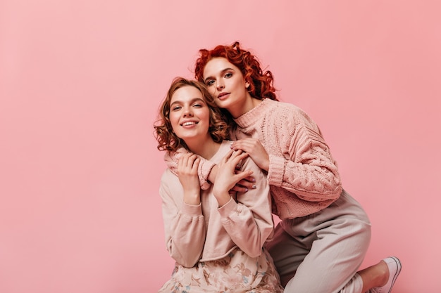 Magníficas chicas en ropa de moda posando sobre fondo rosa. Foto de estudio de dos amigos abrazándose y mirando a la cámara.