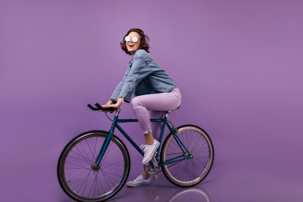 Magnífico ciclista femenino posando en traje de mezclilla Chica elegante riendo sentada en bicicleta