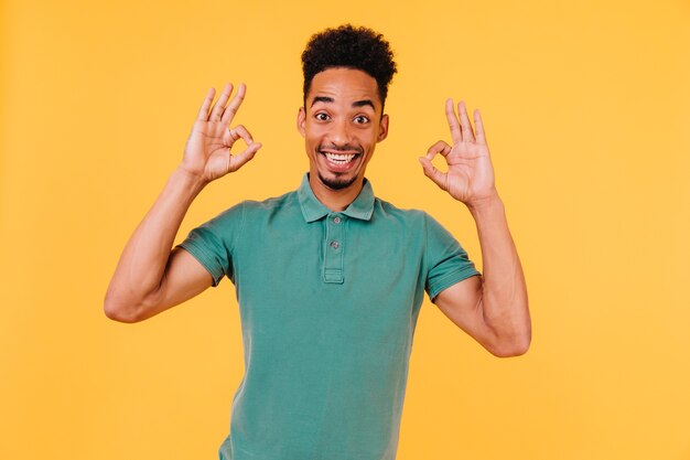 Magnífico modelo masculino en camiseta verde posando con signo bien. Filmación en interiores de hombre africano dichoso expresando emociones positivas.