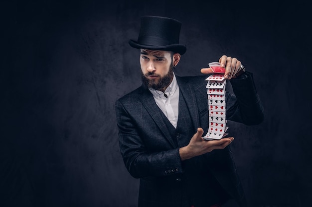 Un mago barbudo con traje negro y sombrero de copa, mostrando trucos con cartas en un fondo oscuro.