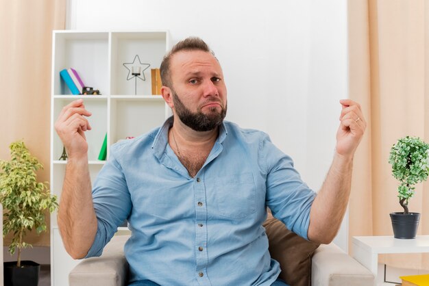 Malestar hombre eslavo adulto se sienta en un sillón gesticulando dinero signo de mano con las dos manos mirando a la cámara dentro de la sala de estar