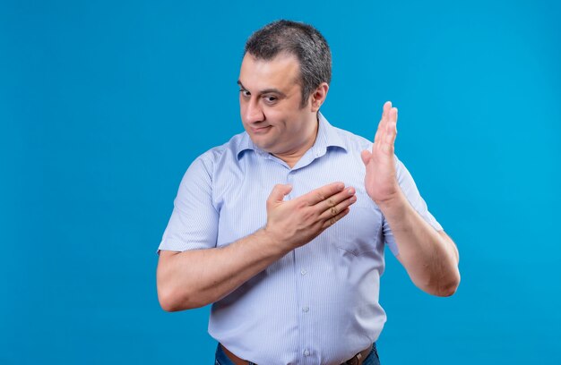 Malestar hombre de mediana edad en camisa azul a rayas mostrando con el dedo índice en el centro de la palma sobre un fondo azul.