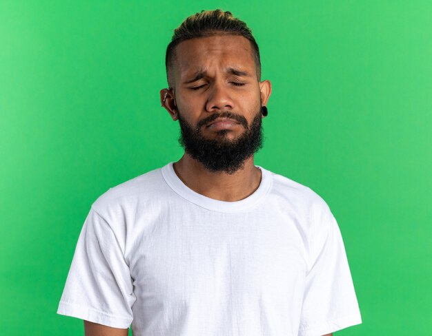 Malestar joven afroamericano en camiseta blanca con expresión triste frunciendo los labios con los ojos cerrados