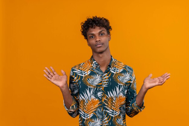 Malestar joven guapo de piel oscura con cabello rizado en hojas camisa estampada abriendo sus manos sobre un fondo naranja