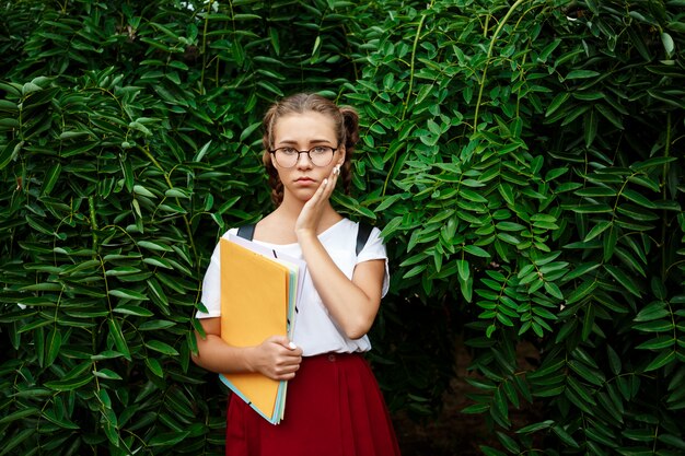 Malestar joven hermosa estudiante en vasos con carpetas al aire libre.