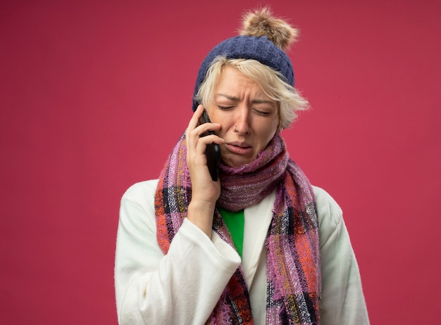 Foto gratuita malestar mujer enferma insalubre con pelo corto en bufanda caliente y sombrero malestar llorando mientras habla por teléfono móvil parado sobre pared rosa