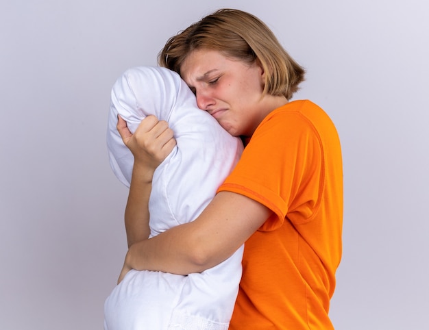 Malestar mujer joven insalubre en camiseta naranja sosteniendo la almohada sintiéndose enferma sufriendo de gripe llorando mucho de pie sobre la pared blanca