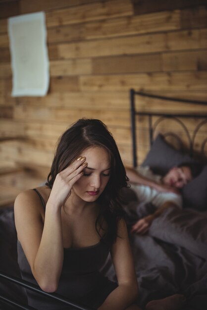 Malestar mujer sentada en una cama mientras el hombre durmiendo en segundo plano.