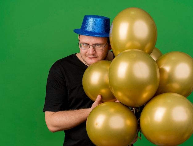 Malvado hombre eslavo adulto alegre en gafas ópticas con gorro de fiesta azul sostiene globos de helio