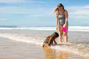 Foto gratuita mamá alegre e hija de pie hasta los tobillos en agua de mar y arena húmeda, recogiendo conchas en balde