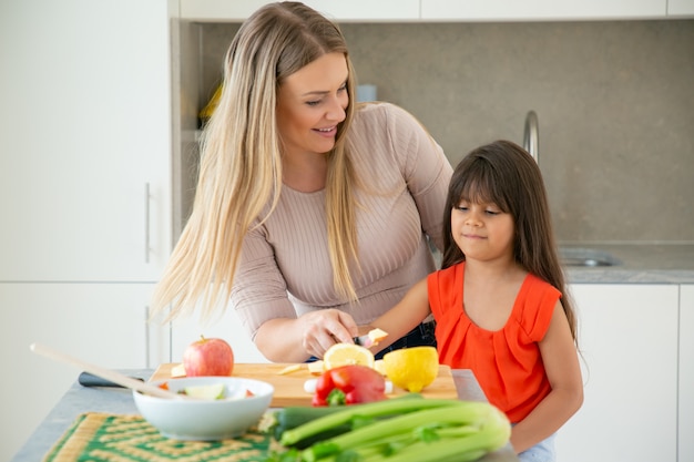 Mamá alegre enseñando a su hija a cocinar ensalada. Niña y su madre cortando verduras frescas en la mesa de la cocina. Concepto de cocina familiar