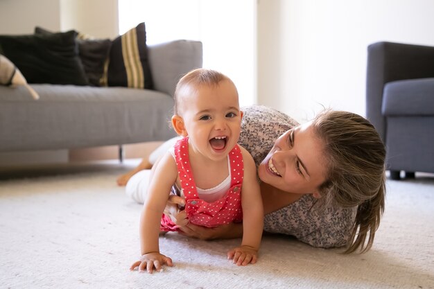 Mamá alegre y niña acostada sobre una alfombra en la sala de estar. Mamá rubia feliz jugando con hija alegre en casa y sonriendo. Lindo bebé riendo con la boca abierta. Concepto de maternidad y fin de semana.