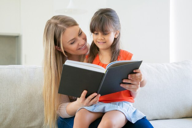 Mamá alegre y su libro de lectura de niña de cabello negro juntos en casa.