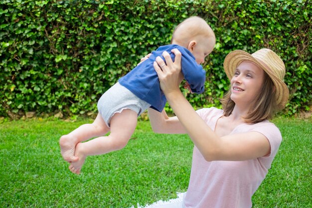 Mamá atractiva con sombrero jugando con recién nacido, sonriendo y mirándolo. Bebé pelirrojo en camisa azul en manos de la madre