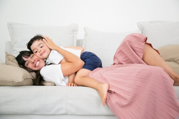 Mamá bastante joven acostada en el sofá y abrazando a un hijo lindo con amor.