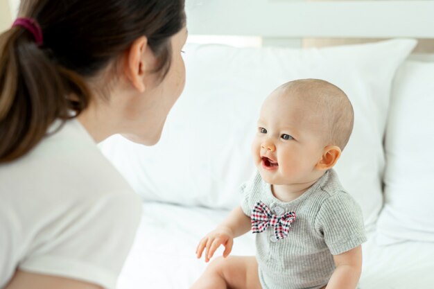 Mamá y bebé felizmente se burlan mutuamente en una cama blanca.