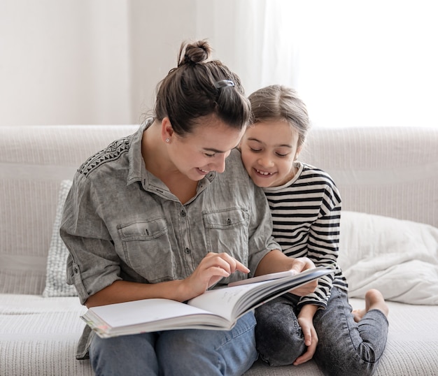 Mamá e hija alegres están descansando en casa, leyendo un libro juntos