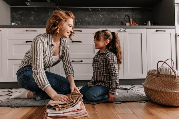 Mamá e hija con un atuendo similar charlan amablemente mientras están sentadas en el piso de la cocina y doblan la ropa lavada.