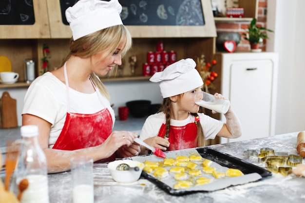 Foto gratuita mamá e hija se divierten preparando galletas con leche en una mesa en una cocina acogedora