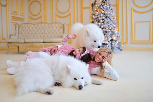 Mamá e hija juegan con perros Samoyedo antes de un árbol de Navidad