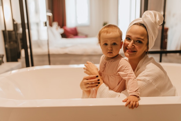 Mamá e hija pequeña miran a la cámara con una sonrisa mientras están sentadas en el baño blanco.