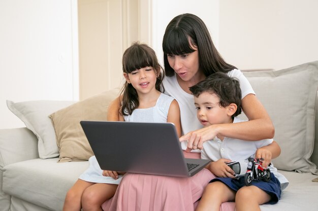 Mamá emocionada alegre abrazando a niños felices y apuntando a la pantalla del portátil. Familia sentada en el sofá en casa y viendo una película.