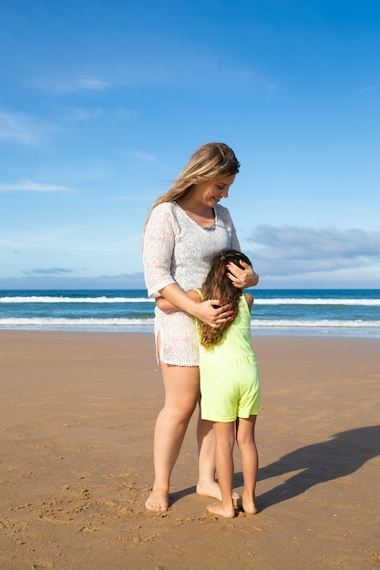 Mamá feliz y dulce hijita en ropa de verano abrazándose mientras está de pie en la playa del océano