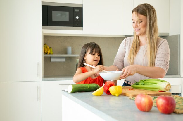 Mamá feliz enseñando a su linda hija a cocinar verduras. Niña ayudando a la madre a tirar la ensalada en la cocina. Concepto de cocina familiar