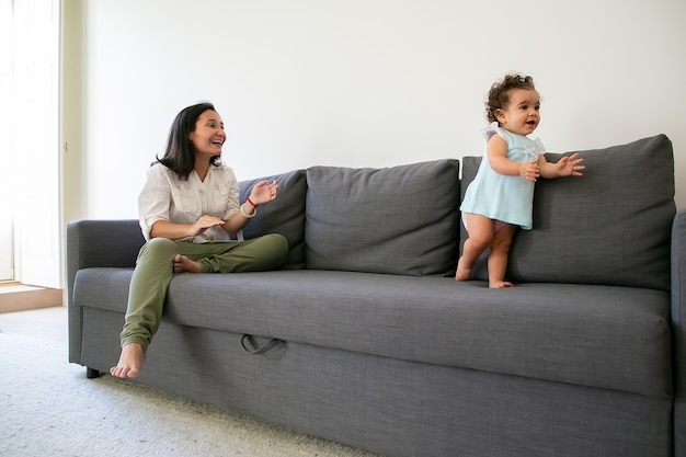 Mamá feliz viendo a la niña dando sus primeros pasos en el sofá. Longitud total. Concepto de paternidad e infancia