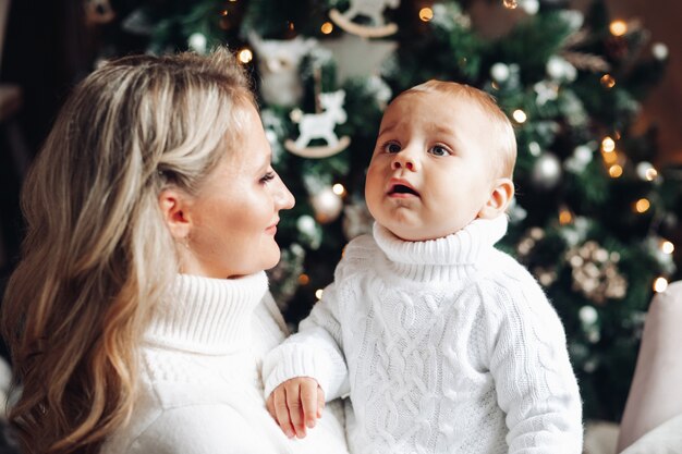 Mamá hermosa se divierte mucho con su bebé cerca del árbol de Navidad en casa