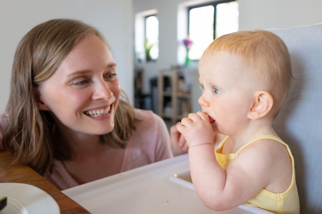 Mamá joven alegre viendo a la hija mordiendo un trozo de comida. Fotografía de cerca. Concepto de nutrición o cuidado infantil