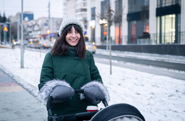 Mamá joven elegante con una taza de café en un paseo con un cochecito de bebé en invierno