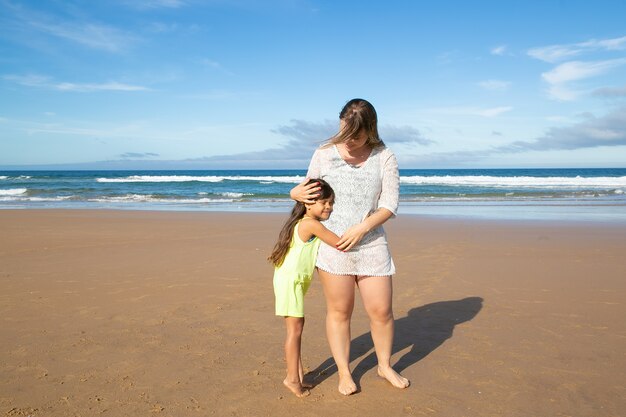 Mamá joven y linda chica de pelo negro abrazándose mientras está de pie en la playa del océano