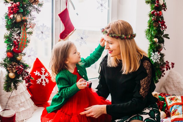 Mamá juega con hija pequeña antes de una ventana brillante decorada para Navidad