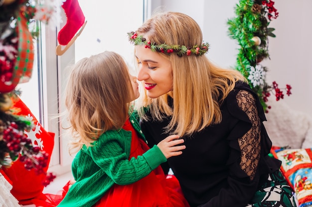 Mamá juega con hija pequeña antes de una ventana brillante decorada para Navidad