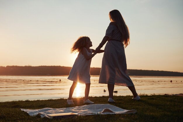 Mamá juega con su bebé de vacaciones cerca del océano, siluetas al atardecer