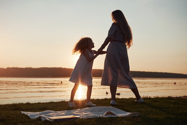 Mamá juega con su bebé de vacaciones cerca del océano, siluetas al atardecer