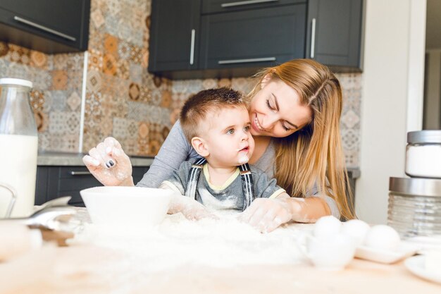 Mamá jugando con niño en la cocina. La cocina está hecha en colores oscuros y estilo rústico. El niño está cubierto de harina y se ve gracioso. La mesa está hecha de madera clara. Licuadora, leche, huevos, frascos de pie sobre la mesa.