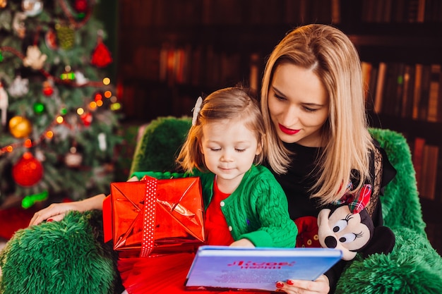 Mamá lee un libro con su pequeña hija sentada en la silla antes del árbol de Navidad