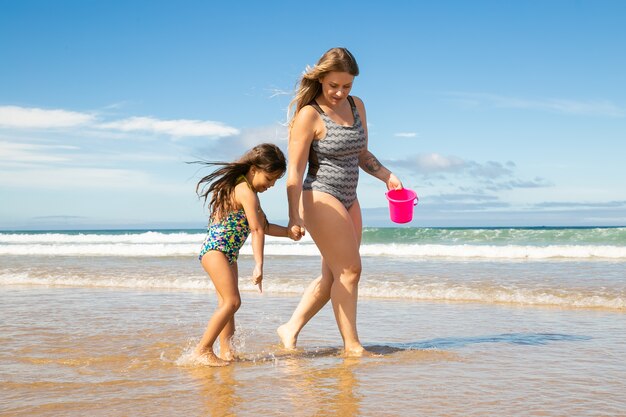 Mamá y niña caminando hasta los tobillos en agua de mar y arena húmeda, recogiendo conchas en balde
